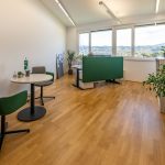Helles, modernes Büro mit bodentiefen Fenstern, grünen Trennwänden und Designmöbeln, ideal für kreatives Arbeiten.