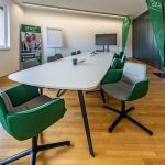 Alt Text: Konferenzraum mit eleganten grünen und grauen Bürostühlen, Whiteboard, Fernseher und Nachhaltigkeitsbanner.