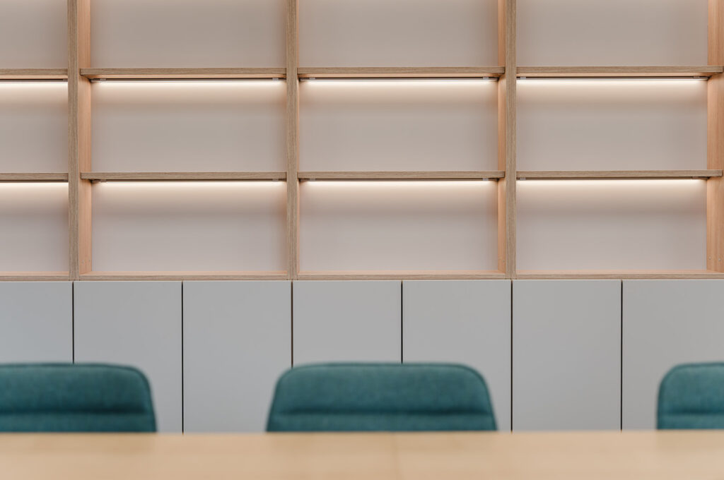 Empty light-wood shelving unit with integrated warm lighting above gray cabinets and teal chairs in the foreground, in a minimalist meeting area.