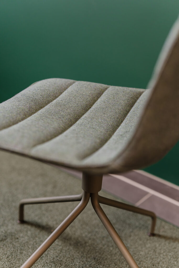 Close-up of a beige upholstered office chair seat with metal legs, against a green wall.