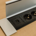 Desk-mounted power module with two European-style sockets and a USB-C port under a metal lid on a wood surface