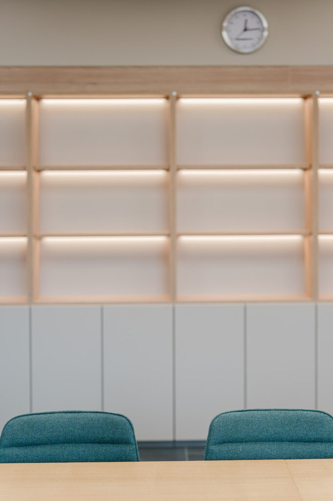Empty meeting room with a wooden table, teal chairs, and a wall of beige shelving under a round clock.