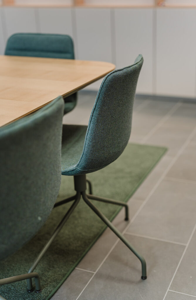 Teal upholstered chair at a wooden conference table in a modern meeting room with a green rug nearby.