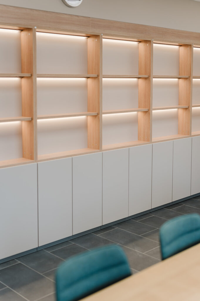 Row of wooden open cubby shelves with integrated lighting above gray cabinets along a tiled floor in a modern workspace.