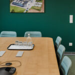 Conference room with a long wooden table, blue chairs, and a tray of glasses with a bottle on green wall backdrop.