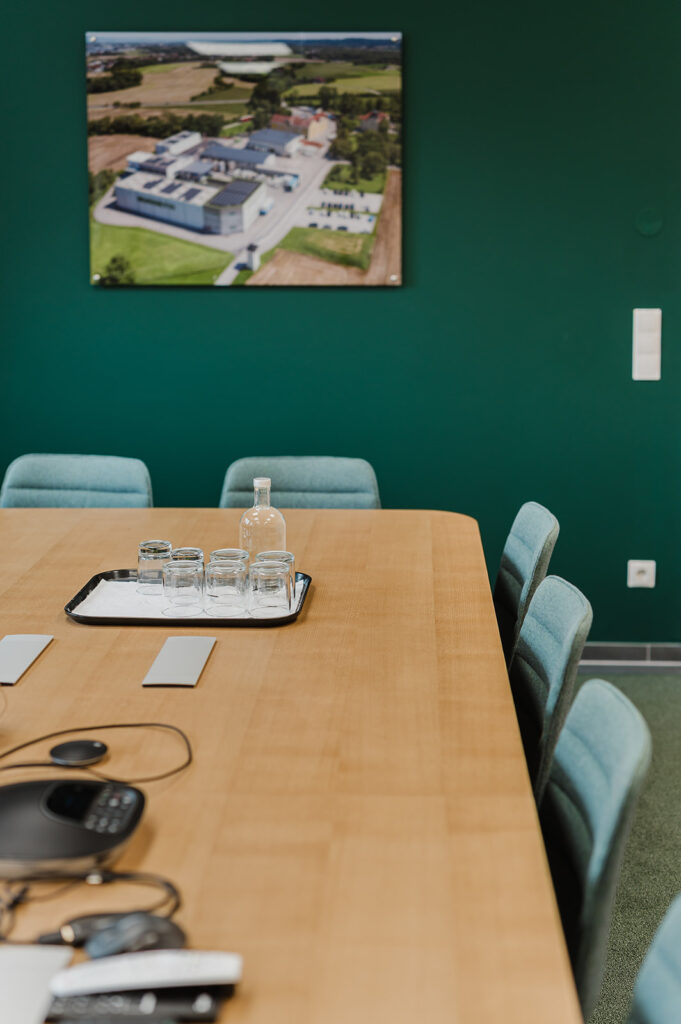 Conference room with a long wooden table, blue chairs, and a tray of glasses with a bottle on green wall backdrop.