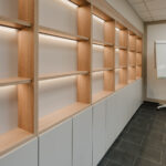 Empty wooden display shelves with integrated lighting running along a wall above gray cabinets.