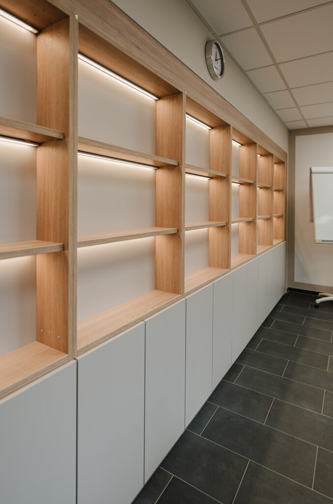 Empty wooden display shelves with integrated lighting running along a wall above gray cabinets.