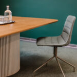 Modern meeting room corner: round-backed gray chair beside a wooden table with a glass water bottle and glasses on a tray, teal wall in background.