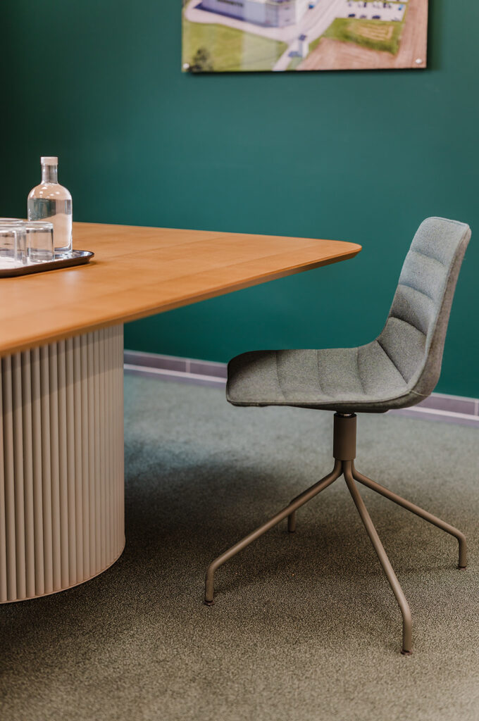 Modern meeting room corner: round-backed gray chair beside a wooden table with a glass water bottle and glasses on a tray, teal wall in background.