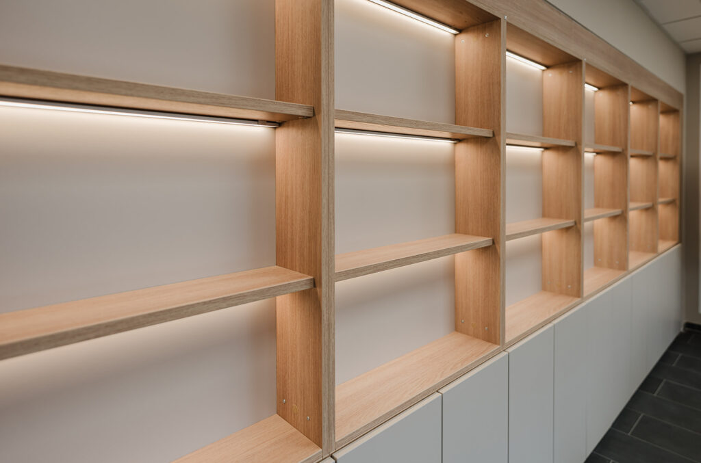 Row of empty wooden display shelves with built-in warm LED lighting along a light wall, viewed from an angled perspective.