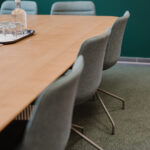 Wooden conference table with a glass water bottle and several glasses on a tray, surrounded by grey chairs in a meeting room with a dark green wall.