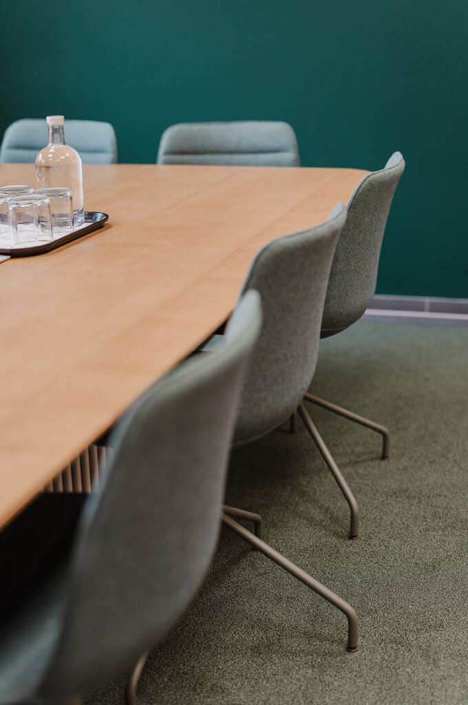 Wooden conference table with a glass water bottle and several glasses on a tray, surrounded by grey chairs in a meeting room with a dark green wall.