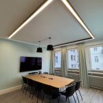 Modern conference room with a long wooden table, black chairs, wall TV, and a large triangular LED ceiling light arrangement above.
