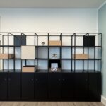 Open, modular black shelving unit with cubby holes and wood accents against a pale blue wall in a showroom setting.