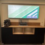 Illuminated display sign with green diagonal stripes and the EICHER logo, mounted on a beige wall above a wooden cabinet.