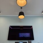 Meeting room with a large wall-mounted flat-screen monitor, two black pendant lights, and a wooden conference table with phones and accessories.