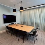Conference room with a long wooden table, black chairs, wall-mounted TV, and two black pendant lights. Pale blue walls and light curtains in the background.