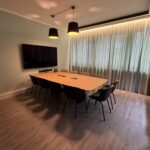 A conference room with a long wooden table surrounded by black chairs, a wall-mounted TV, and warm pendant lights above.