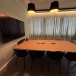 Conference room with a long wooden table, black chairs, and a wall-mounted flat-screen TV; sheer curtains behind.